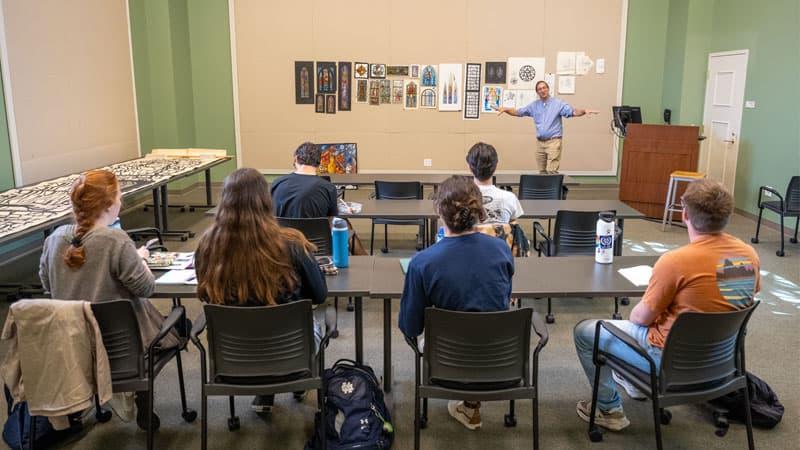 Professor Stephen Hartley stands at the front of a classroom discussing stained glass renderings.