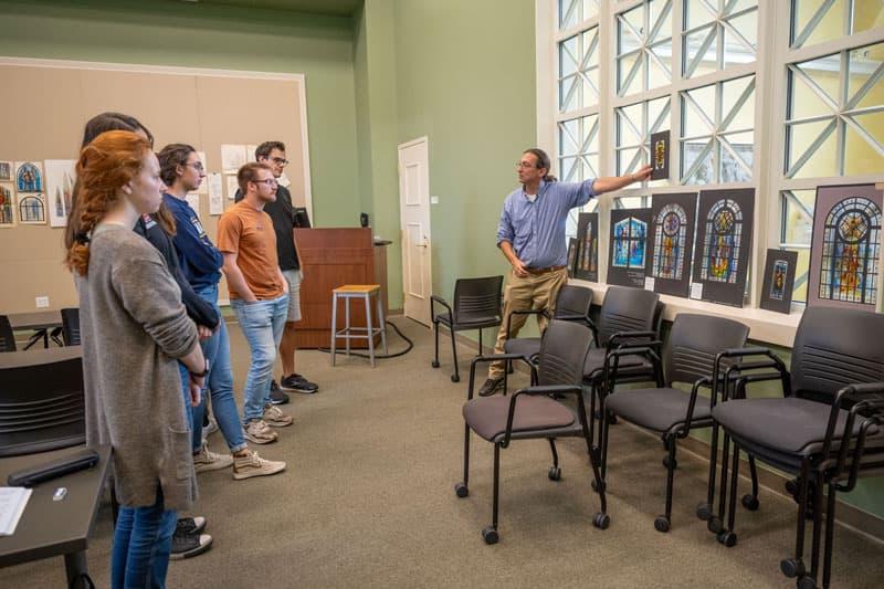 Professor Stephen Hartley discusses stained glass drawings displayed on windows.